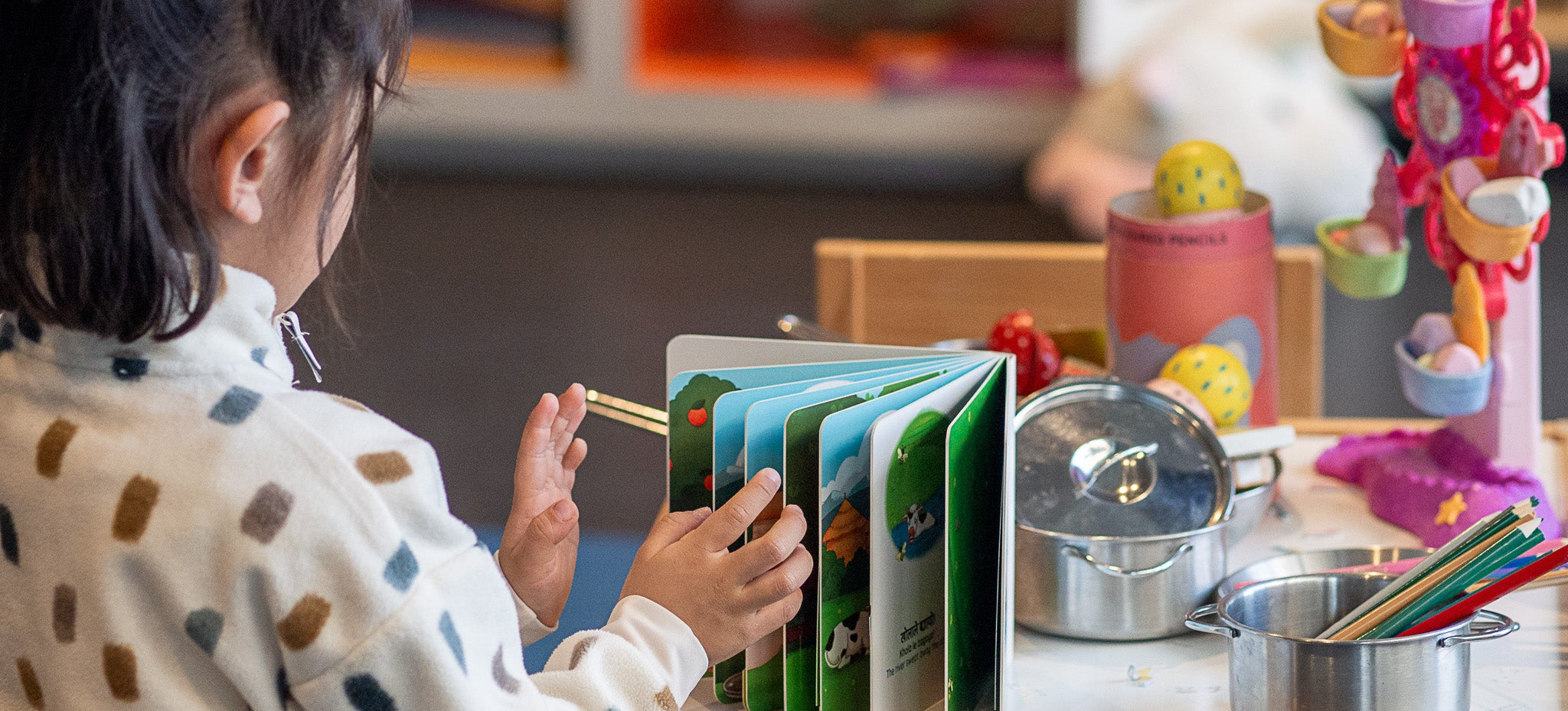 Child interacting with a colorful picture book in Nepali/English at a table with various other toys